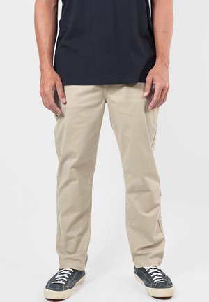 Homme portant un pantalon beige, un t-shirt noir et des baskets noires avec des lacets blancs, debout devant un fond blanc uni.