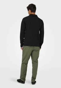 Polo-shirt noir à manches longues avec col, associé à un pantalon vert olive. L'ensemble présente des détails minimaux et une coupe décontractée.