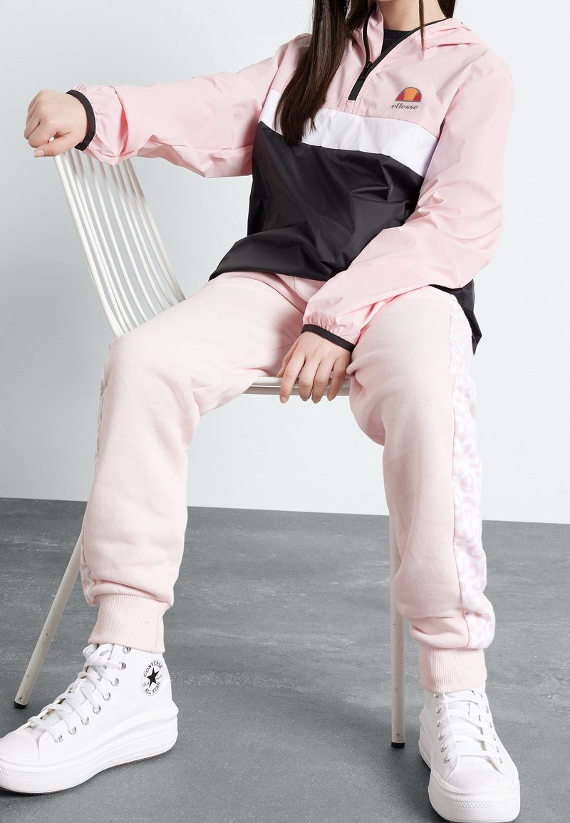 Personne assise sur une chaise blanche portant un pantalon de jogging rose, une veste zippée rose et noire et des baskets montantes blanches Converse.