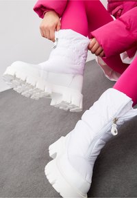 Bílé zateplené boty s tlustou, strukturovanou podešví a bočním zipem. Materiál je lesklý s prošívaným vzorem, což vytváří elegantní moderní vzhled.