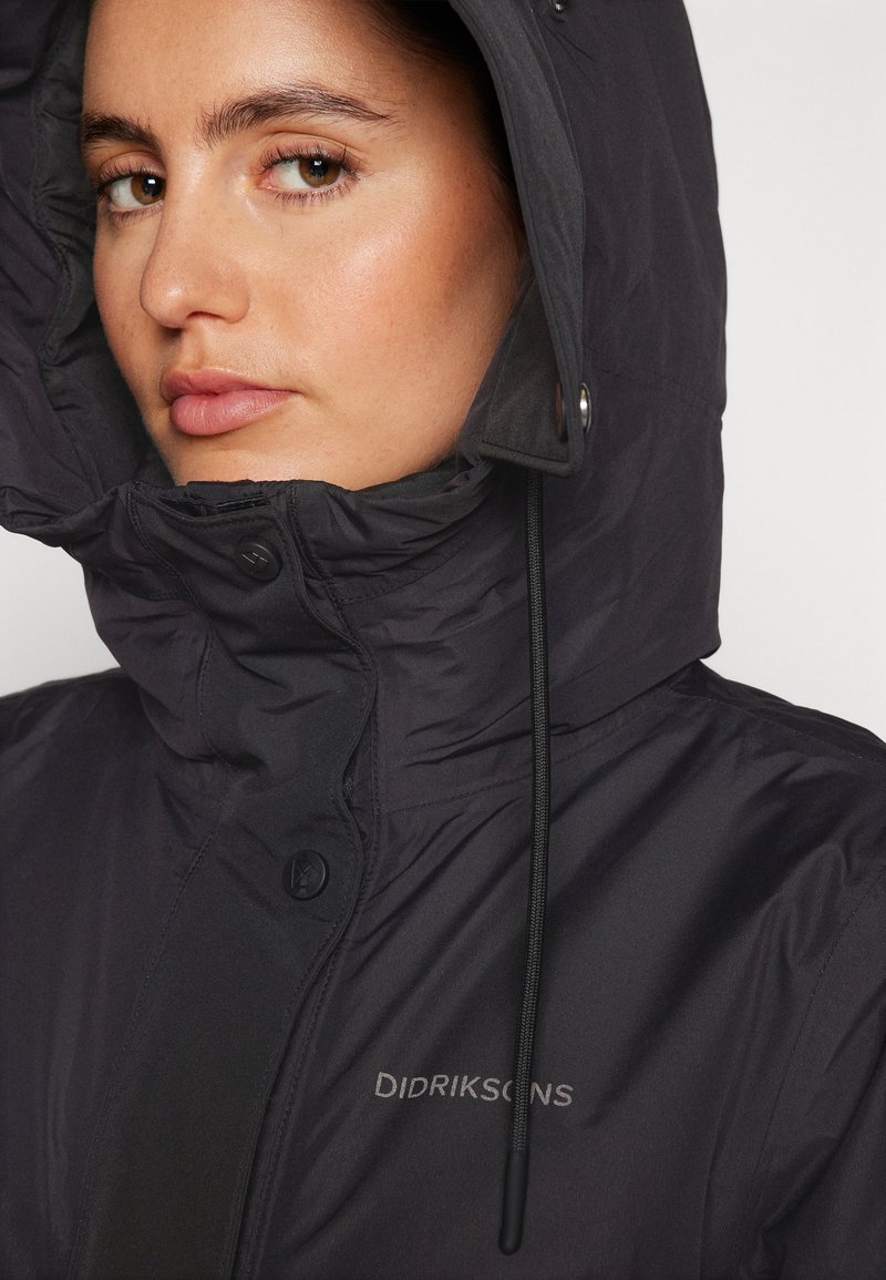 Schwarze, isolierte Jacke mit hohem Kragen, Kapuze, Druckknöpfen und Kordelzug. Branding ist auf der Vorderseite sichtbar. Glatte Textur.