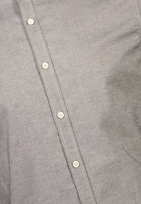 Camicia grigia chiara con cinque bottoni bianchi visibili lungo il centro e cuciture visibili lungo la patta dei bottoni.