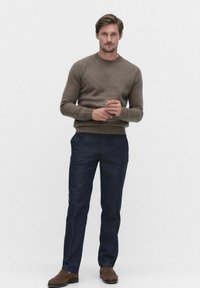 Ruskea villaneule, jossa on pyöreä kaula-aukko, yhdistetty tummaan siniseen housuihin. Malli seisoo kädet yhdessä, jalassa ruskeat kengät. Yksinkertainen muotoilu.