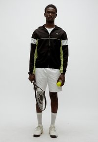 Črna jakna z zadrgo z zelenimi in belimi poudarki, bele kratke hlače, drži rumeno teniško žogico in lopar, stoji na nevtralnem ozadju.