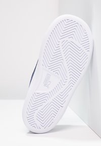 Suela de goma blanca con un patrón de tracción en zigzag, que presenta un logo. Material superior oscuro no visible, apoyándose sobre una superficie clara.