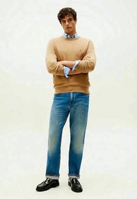 Beige knit sweater, light blue collared shirt underneath, faded blue jeans, and black glossy shoes. Standing with arms crossed against a light background.
