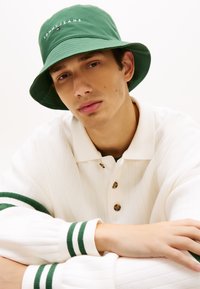 Πράσινο κάσκα με κεντημένο λογότυπο "TOMMY JEANS"· λευκό πλεκτό πουλόβερ με πράσινες ρίγες στις μανσέτες· λεπτομέρειες με κουμπιά στο γιακά.