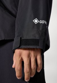 Sort vandtæt jakke med velcro-manchet, prydet med et hvidt "GORE-TEX" logo, glat tekstur og en tætsiddende pasform, der er synlig på ærmet.