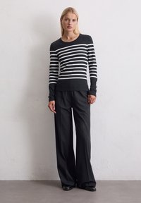 Kvinne står mot en strukturert hvit vegg iført en svart og hvit stripete langermet topp, vide svarte bukser og svarte sko.