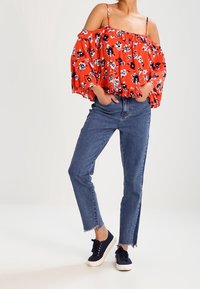 Femme portant un jean bleu avec des ourlets bruts, des baskets bleu marine, et un chemisier rouge à fleurs décolleté sur les épaules avec des manches à volants et des bretelles.