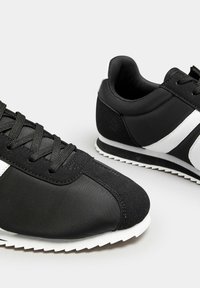 Black athletic shoes made of synthetic material, featuring a textured upper, white side stripes, and a rubber sole with a zigzag pattern.