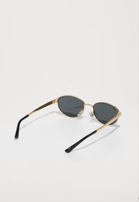 Ochelari de soare tip cat-eye din metal auriu cu lentile gri, având vârfuri de temple din plastic negru și accesorii pentru nas pentru confort.