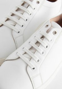 White sneakers featuring a smooth leather upper, round toe, six pairs of eyelets, and flat laces. Minimalist design with white stitching.