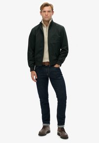 Chaqueta bomber verde oscura sobre un suéter crema, combinada con jeans de mezclilla oscuros y botas marrones con cordones. Presenta un cierre en la parte delantera y puños acanalados.