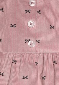 Pink corduroy dress featuring vertical stripes, black bow patterns, and three shiny buttons with a gathered waistline detail.