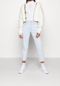 Chaqueta blanca corta sobre una blusa sin mangas blanca, combinada con unos vaqueros de tiro alto azul claro y zapatillas blancas. Diseño sencillo, sin patrones.