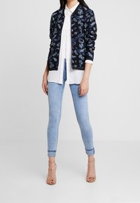 Marinblå bomberjacka med paisleymönster, bärs över en vit skjorta med knappar, i kombination med ljusblå skinny jeans och beige sandaler med remmar.