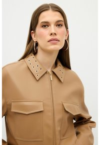 Tan faux læderjakke med lynlås, stor krave med øje-huls detaljer og to frontlommer. Glat tekstur og moderne design.