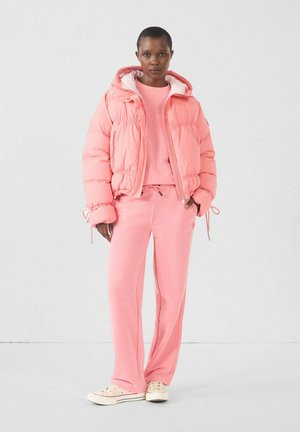 Roze pufferjack met een capuchon en strikkende manchetten, gecombineerd met een bijpassende roze trui en joggingsbroek, gedragen met beige sneakers.