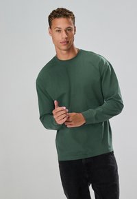 Green long-sleeve t-shirt made of cotton, featuring a round neckline and ribbed cuffs, paired with black pants. Smooth texture.