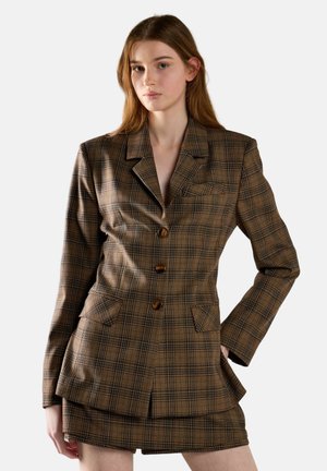 Joven mujer con blazer marrón de cuadros y falda a juego, de pie con una mano en el bolsillo frente a un fondo liso.