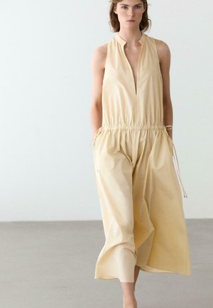 Kvinde iført ærmeløs beige jumpsuit med snøre i taljen og vide ben, stående foran en ensfarvet lys baggrund.