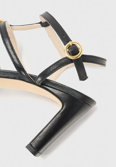 Sandalias de tacón alto negras con tiras entrelazadas, acento en anillo dorado, textura de cuero suave y diseño de tacón grueso.