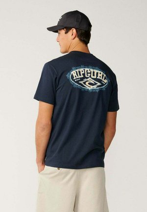 Man met een zwarte pet, donkerblauw Rip Curl T-shirt met logo op de rug en lichtbeige short, zijwaarts staand met handen in de zakken.