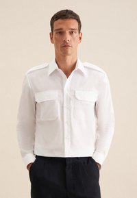 Witte button-up shirt met twee borstzakken, gestructureerde schouders en een gladde textuur. Vergezeld van donkere broeken.
