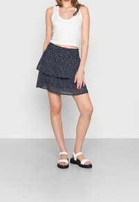 Navy blue, layered wrap skirt with white polka dots; paired with a fitted, white sleeveless top and white sandals with black soles.