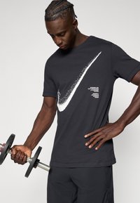 Μαύρο t-shirt Nike με μεγάλο λευκό λογότυπο Swoosh, κοντά μανίκια, στρογγυλή λαιμόκοψη και λεπτομέρειες κειμένου στο μπροστινό μέρος, φορεμένο ενώ σηκώνει αλτήρα.
