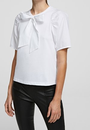 Vrouw draagt een witte blouse met korte mouwen, pofschouders en een grote strik bij de halslijn, gecombineerd met zwarte, nauwsluitende broek.