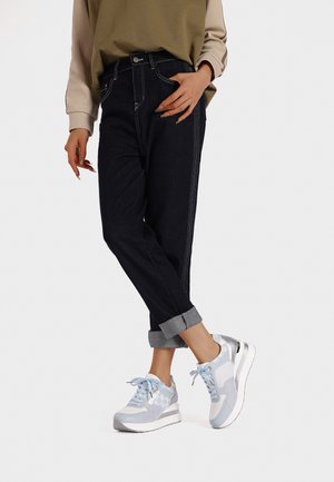 Jeans alti blu scuro con cuciture bianche a contrasto, risvoltini sulle caviglie, abbinati a sneaker grigie chiare e blu con dettagli in rete.
