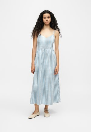 Frau steht in einem blau-weißen, ärmellosen Kleid mit Spaghettiträgern und beigen Ballerinas frontal vor einem schlichten Hintergrund.