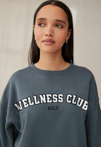 Šedá mikina s okrúhlym výstrihom, s bielym texturálnym nápisom "WELLNESS CLUB" a čiernym nápisom "SELF." v uvoľnenom strihu a mäkkej látke.
