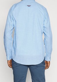 Jasnoniebieska, kratkowana koszula z długim rękawem, zapinana na guziki przy mankietach, o zaokrąglonym dole. Na karku z tyłu znajduje się detal z kontrastowego materiału.