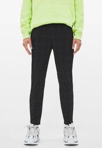 Man med slim-fit svarta byxor med subtilt rutig mönster, vita sneakers och ljusgrön hoodie, står mot vit bakgrund.