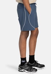 Dunkelblaue Sports Shorts mit weißen Paspeln, elastischem Bund und seitlichen Taschen, kombiniert mit schwarzen Sneakers und Crew-Socken.