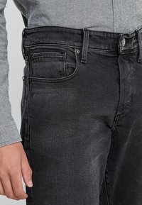 Vaqueros de mezclilla negra con corte recto, que cuentan con trabillas para cinturón, cinco bolsillos y una textura ligeramente desgastada. Cierre de botón y cremallera.