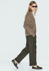 Maglione a maglia marrone, pantaloni su misura verde oliva e scarpe slip-on scure. Il modello indossa occhiali da sole scuri e cammina su uno sfondo chiaro.
