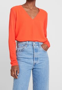 Top de manga larga con escote en V en tejido naranja brillante, combinado con vaqueros de tiro alto en azul claro que tienen una bragueta de botones y bolsillos frontales.