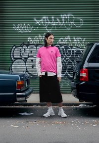 Camiseta rosa de manga corta sobre una camisa blanca de rayas con mangas largas, pantalones culottes negros y zapatillas blancas. De fondo, un muro verde con grafiti.