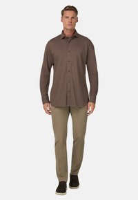 Hnědá košile s dlouhým rukávem, zapínací na knoflíky, s límečkem, spárovaná se béžovými kalhotami a tmavými botami. Materiál vypadá jemně a hladce.