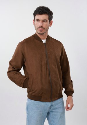 Mees, kes kannab pruuni luku ja pealiskattega bomber-jopet ning helesiniseid teksaseid, poseerib ühe käega taskus, neutraalse taustaga.