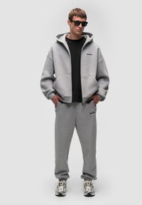 Světle šedá zipová mikina s kapsami a černým logem, spárovaná s odpovídajícími zúženými tepláky, nošená přes černé tričko a tenisky.