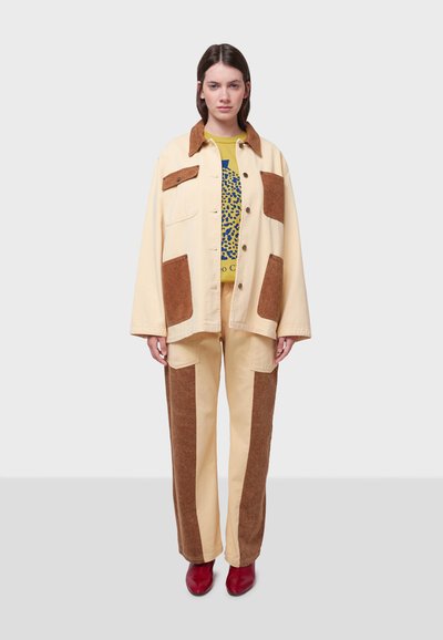 Beige kavaj med bruna manchesterlappar, parat med beige byxor med bruna sidopanler. Modellen bär röda skor och en t-shirt med grafiskt tryck.