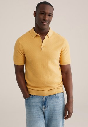 Geel poloshirt met korte mouwen en een gestructureerd patroon, voorzien van een kraag en een drieknops sluiting, gecombineerd met lichtblauwe spijkerbroek.