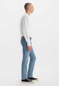 Man in een wit overhemd met lange mouwen, lichte blauwe jeans en zwarte sneakers staat in profiel tegen een effen witte achtergrond.