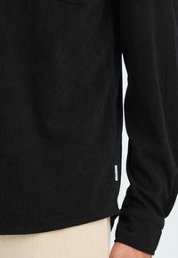 Must musträp shirt, millel on ribid, pikad varrukad ja külgmised lõhed. Sisaldab väikest logo sildi alumisel vasakul äärel.