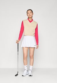 Golfoutfit met een beige mouwloos vest over een roze langemouwhemd, gecombineerd met een witte rok en witte Nike-schoenen. Houdt een golfclub vast.